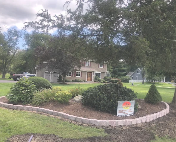 Red brick colonial house with manicured landscaping, curved flower bed border, and realtor sign in front yard